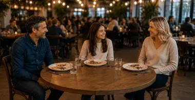 Trzy osoby rozmawiają przy okrągłym stole w restauracji, z talerzami i szklankami.
